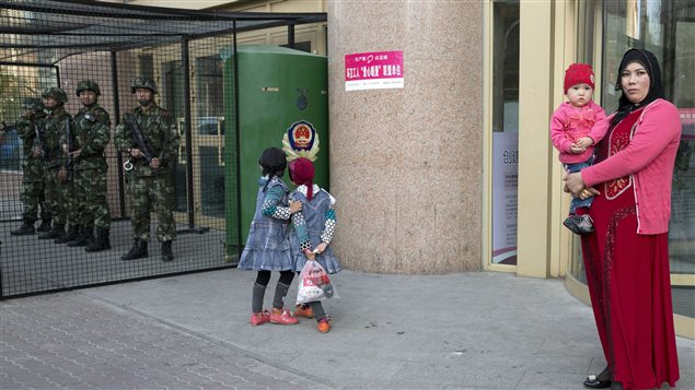 A Uighur woman carries a toddler as children play near a cage protecting heavily armed Chinese policemen in Xinjiang on May 1, 2014. Government directives prohibited names like Muhammad, Jihad, and Islam for children in the mainly Muslim region.